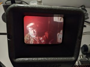 Still from 16mm film of man with black and white makeup wearing black google and red military style jacket